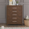 The Child Craft Kieran 5-Drawer Chest in Toasted Chestnut stands against a gray paneled wall. Its five drawers have pink handles, and on top are books, décor, and a plant. A teddy bear sits on a basket nearby, next to a rug.