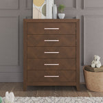 The Child Craft Kieran 5-Drawer Chest in Toasted Chestnut stands against a gray paneled wall. Its five drawers have pink handles, and on top are books, décor, and a plant. A teddy bear sits on a basket nearby, next to a rug.