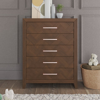The Child Craft Kieran 5-Drawer Chest in Toasted Chestnut stands against a gray paneled wall. Its five drawers have pink handles, and on top are books, décor, and a plant. A teddy bear sits on a basket nearby, next to a rug.