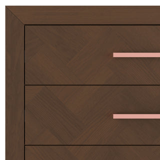Close-up of the Kieran 5-Drawer Chest by Child Craft in Toasted Chestnut, showing a herringbone pattern on the drawer fronts. Two horizontal drawers have straight pink handles, and the wood features a rich, dark brown finish.