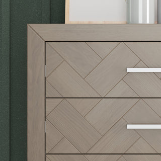 Close-up of the Child Craft Kieran 5-Drawer Chest in Crescent Gray, featuring herringbone-patterned drawers with modern silver handles, topped with a framed picture and vase, set against a dark green textured wall.
