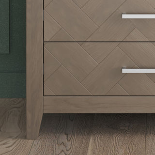 A close-up of the Child Craft Kieran 5-Drawer Chest in Crescent Gray, showing its herringbone pattern and sleek silver handles. The chest sits on a wooden floor beside a dark green wall.