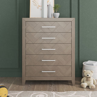 The Child Craft Kieran 5-Drawer Chest in Crescent Gray stands against a green wall, topped with decor, art, and a small plant. Nearby, a teddy bear and toys rest on the wooden floor.