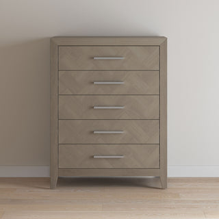 The Child Craft Kieran 5-Drawer Chest in Crescent Gray, with sleek silver handles, stands against a plain off-white wall on a light wood floor.