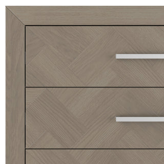 Close-up of the upper left corner of the Child Craft Kieran 5-Drawer Chest in Crescent Gray, showing a light wood finish with a herringbone pattern and two horizontal metal handles on the drawers.