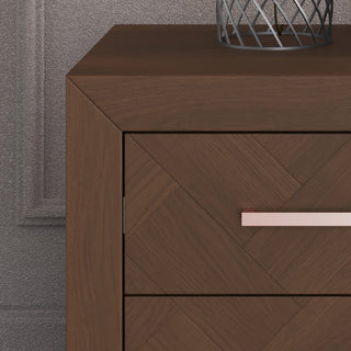 A close-up of the Child Craft Kieran Nightstand in Toasted Chestnut, featuring a geometric drawer pattern, metallic handle, and baby-safe finishes, with a decorative object on top and set against a textured wall.