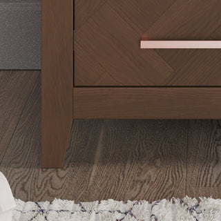 Close-up of a Child Craft Kieran Nightstand in Toasted Chestnut, featuring a copper handle and sturdy leg on a textured rug and wood floor—an ideal nursery nightstand with baby-safe finishes. White fabric object partially visible bottom left.