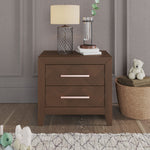 The Child Craft Kieran Nightstand in Toasted Chestnut with two drawers provides ample bedside storage against a gray paneled wall, topped with a lamp, books, and water. A nearby toy basket makes it ideal for a baby’s nursery.