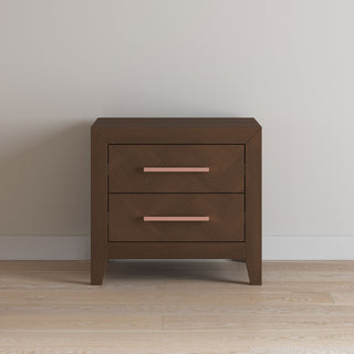 The Child Craft Kieran Nightstand in Toasted Chestnut, featuring two drawers with pink handles and baby-safe finishes, stands against a plain light-colored wall on a hardwood floor.