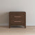 The Child Craft Kieran Nightstand in Toasted Chestnut, featuring two drawers with pink handles and baby-safe finishes, stands against a plain light-colored wall on a hardwood floor.