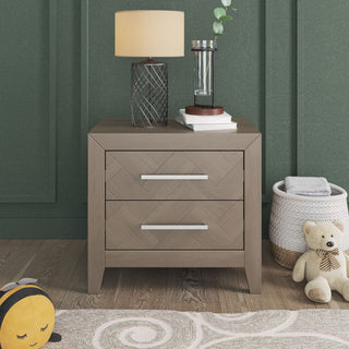 The Child Craft Kieran Nightstand in Crescent Gray stands against a green wall with a lamp, plant, and books on top. Nearby, a plush bee, teddy bear, basket with blanket, and part of a patterned rug are visible.