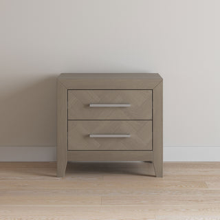 The Child Craft Kieran Nightstand in Crescent Gray features two drawers with metal handles and angled legs, set against a light wall and wooden floor.