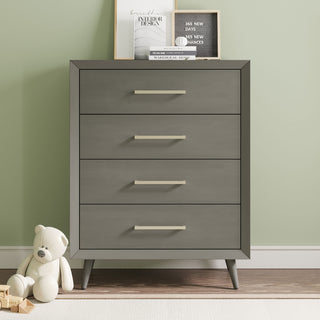 The Child Craft Cranbrook 4 Drawer Chest in Lunar Gray stands against a light green wall, topped with framed photos and a letterboard. A white teddy bear and wooden blocks rest on the floor next to the dresser.