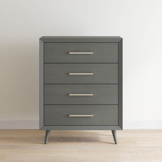 The Child Craft Cranbrook 4 Drawer Chest in Lunar Gray features sleek metal handles and angled legs, standing stylishly against a white wall on a light wooden floor.