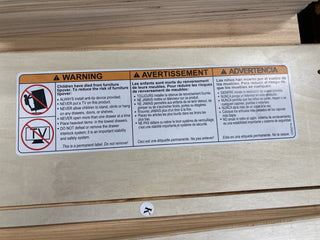 A safety label on the Child Craft Cranbrook 4-in-1 Convertible Crib, Toasted Chestnut, provides tip-over prevention instructions in English, French, and Spanish: do not allow children to climb or open drawers and secure the crib to the wall.