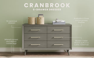 The Child Craft Cranbrook Double Dresser in Lunar Gray stands against a green wall, featuring modern metal handles, angled legs, mitered corners, and spacious drawers—an ideal piece of stylish mid-century modern nursery furniture.