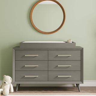 A modern Child Craft dresser in Lunar Gray with six drawers stands by a light green wall. On top is the F01792 Universal Changing Table Topper, with baby items, a round wooden mirror above, and a white teddy bear on the floor to the left.