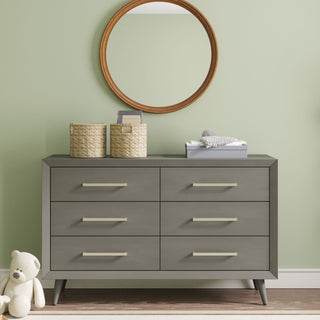A Child Craft Cranbrook Double Dresser in Lunar Gray with six drawers sits against a light green wall. Two woven baskets and folded baby clothes rest on top, with a round wooden-framed mirror above and a white teddy bear on the floor nearby.