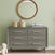 A Child Craft Cranbrook Double Dresser in Lunar Gray with six drawers sits against a light green wall. Two woven baskets and folded baby clothes rest on top, with a round wooden-framed mirror above and a white teddy bear on the floor nearby.