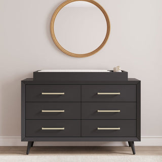 A sleek black dresser with six drawers and brass handles stands against a light wall, topped with the Child Craft F01792 Universal Changing Table Topper in Ebony and a small container. Above it hangs a round mirror with a wooden frame.
