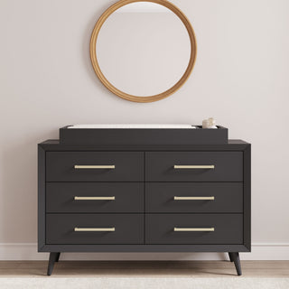 The Child Craft Cranbrook Double Dresser in Ebony, featuring six drawers with gold handles, sits below a round wooden-framed mirror with a white changing pad and container on top, making it a stylish addition to the nursery against a light wall.