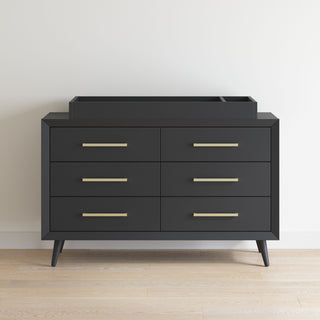 A sleek black six-drawer dresser with gold handles and angled legs stands on a light wood floor, featuring the Child Craft F01792 Universal Changing Table Topper in Ebony for added versatility.