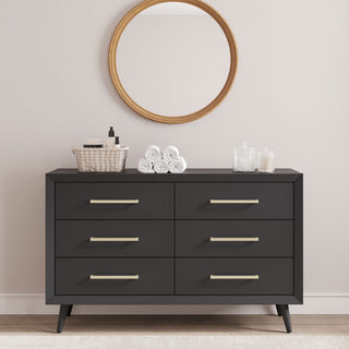 The Cranbrook 2-Piece Nursery Set by Child Craft features a sleek black dresser with six drawers, topped with towels, bottles, and jars. A round wood-framed mirror above completes the Mid Century Modern style against a light wall.