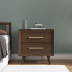 A Child Craft Cranbrook Nightstand in Toasted Chestnut with two drawers and angled legs stands beside a bed, topped with books and decor. A muted green wall, wooden floor, and white rug create a cozy atmosphere.