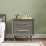 The Child Craft Cranbrook Nightstand in Lunar Gray stands beside a bed, topped with two wooden animal figurines and stacked books. A light green wall and wood floor with a white baseboard complete the scene.
