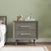 The Child Craft Cranbrook Nightstand in Lunar Gray stands beside a bed, topped with two wooden animal figurines and stacked books. A light green wall and wood floor with a white baseboard complete the scene.