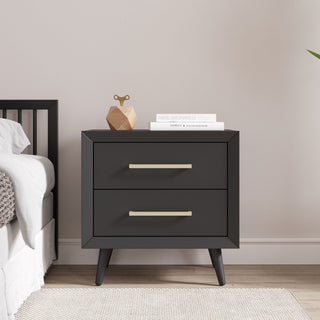The Child Craft Cranbrook Nightstand in Ebony, featuring two drawers with gold handles, is placed by the bed. On top are books, a decorative piece, and a green plant’s leaf; light walls and wood flooring complete the room.