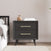The Child Craft Cranbrook Nightstand in Ebony, featuring two drawers with gold handles, is placed by the bed. On top are books, a decorative piece, and a green plant’s leaf; light walls and wood flooring complete the room.