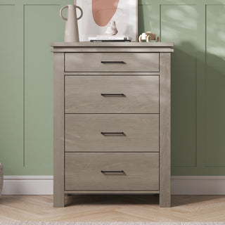The Child Craft Denman 4 Drawer Chest in Crescent Gray stands against a green paneled wall, topped with a ceramic vase, book, and framed abstract art. Sunlight casts shadows over its sleek drawers and black handles.