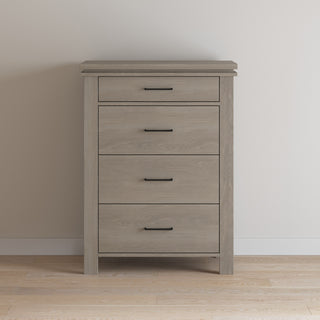 The Child Craft Denman 4 Drawer Chest in Crescent Gray, featuring sleek black handles, stands against a plain white wall on a light wood floor.