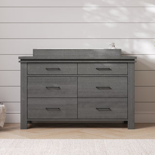 The Child Craft Denman Double Dresser in Midnight Gray, featuring six drawers with horizontal handles, stands against a white shiplap wall. A rectangular sink rests on top, illuminated by sunlight that casts gentle shadows.