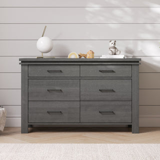 The Child Craft Denman 6-Drawer Dresser in Midnight Grey stands against a white shiplap wall, topped with a round white lamp, decor accents, stacked books, and a toy bear. A wood floor and part of a light rug are also visible.