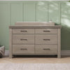 A light wood dresser with six drawers and black handles sits against a green paneled wall. On top is the Child Craft Universal Dressing Kit in Crescent Grey (F01792), with a soap dispenser and basket nearby. Sunlight casts soft shadows across the wall.