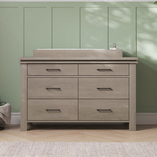 A light wood dresser with six drawers and black handles sits against a green paneled wall. On top is the Child Craft Universal Dressing Kit in Crescent Grey (F01792), with a soap dispenser and basket nearby. Sunlight casts soft shadows across the wall.