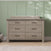 The Child Craft Denman Double Dresser in Crescent Gray features six drawers with dark handles and is placed against a green paneled wall on a light herringbone wood floor, with leaf shadows visible on the wall.