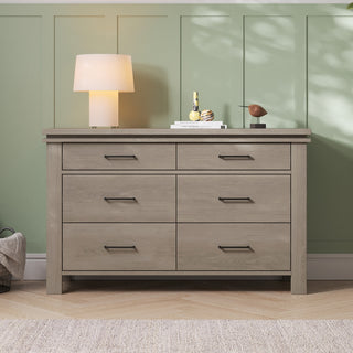 A Child Craft Denman Double Dresser in Crescent Gray with six drawers stands against a green paneled wall. On top sit a lamp, stacked books, decor pieces, and a small sculpture as sunlight casts shadows on the wall and floor.