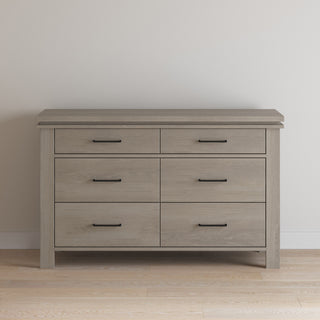 The Child Craft Denman Double Dresser in Crescent Gray, featuring six drawers with black handles, stands against a plain light-colored wall on a wooden floor.