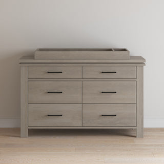 The Child Craft Denman Double Dresser in Crescent Gray, featuring six drawers with black handles and a matching wooden changing tray, stands against a light wall on a wooden floor.