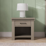 A Child Craft Denman Nightstand in Crescent Gray, featuring a single drawer and open shelf, stands against a green paneled wall with a white cylindrical table lamp on top and is set on a light wood floor.