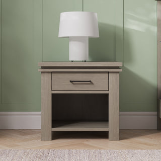 A Child Craft Denman Nightstand in Crescent Gray, featuring a single drawer and open shelf, stands against a green paneled wall with a white cylindrical table lamp on top and is set on a light wood floor.