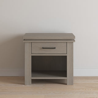 The Child Craft Denman Nightstand in Crescent Gray features a single drawer with a black handle and open storage below, making it ideal for nursery furniture or as a modern accent piece in any room.