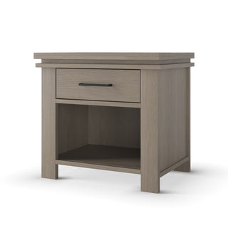 The Child Craft Denman Nightstand in Crescent Gray features a single drawer with black handle and an open lower shelf, ideal for modern bedrooms or nurseries. Shown on a white background.
