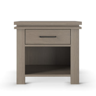 The Child Craft Denman Nightstand, Crescent Gray, is a modern piece featuring a light gray finish, one drawer with a black handle, and an open lower shelf—perfect for nurseries or stylish bedrooms.