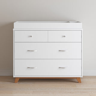 A white double dresser with two small top drawers and two large bottom drawers stands on wood legs with sleek handles. Against a light wall on a wood floor, it’s topped with the Child Craft Legacy Changing Table Topper in Matte White for nursery versatility.