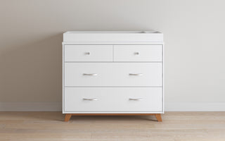 A modern white double dresser with four drawers and round knobs stands on wooden legs against a light wall and hardwood floor, and pairs easily with the Child Craft Legacy Changing Table Topper in Matte White.