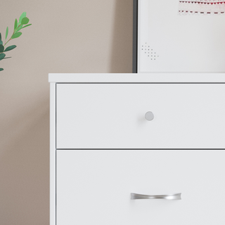 A close-up of the Child Craft Soho 3-Drawer Dresser in White/Natural shows its distinct knobs and handles, placed beside a green plant with a framed picture partially visible on the wall above.